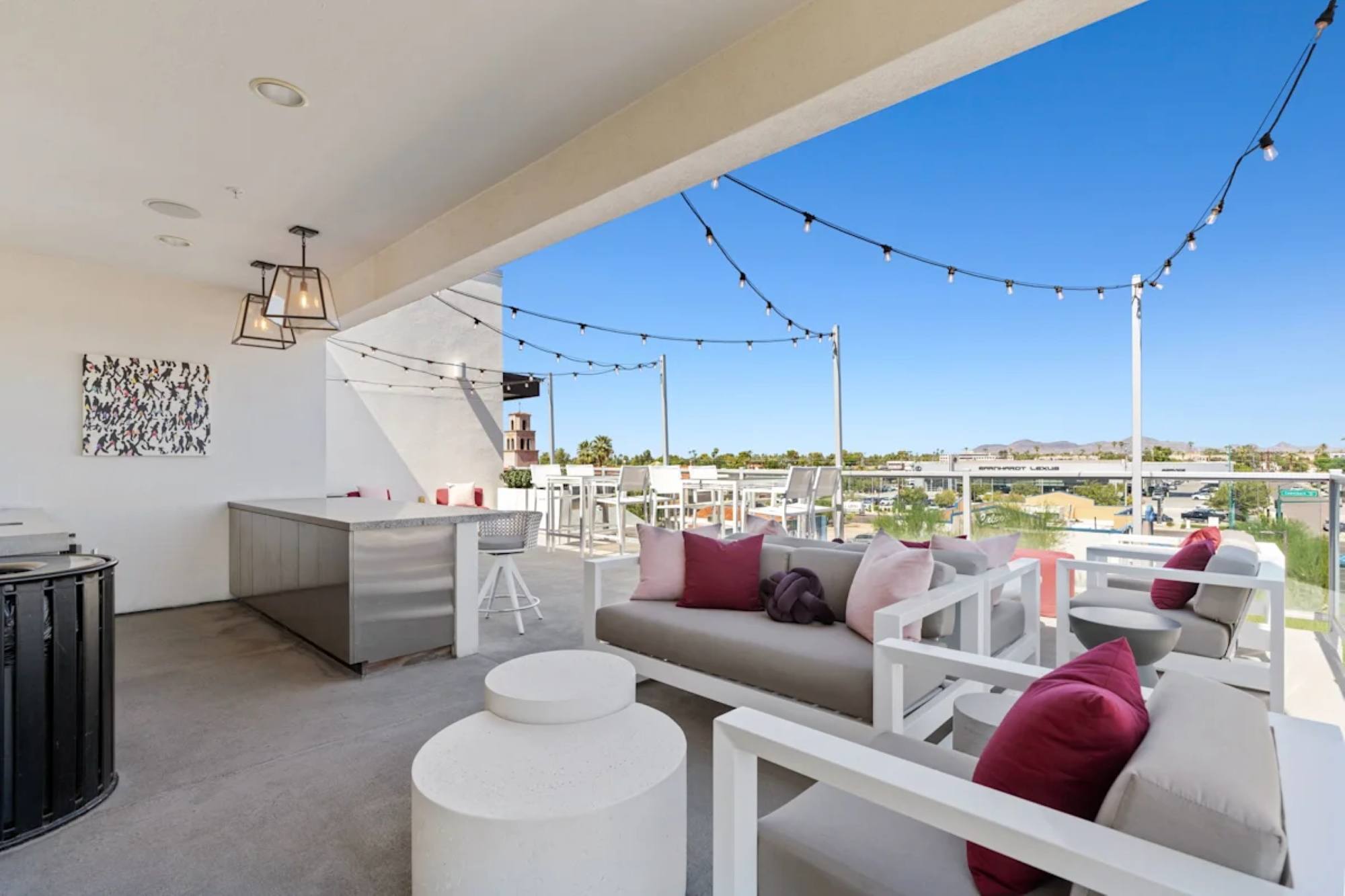 A rooftop lounge area featuring comfortable seating with pink pillows, overhead string lights, and scenic outdoor views.